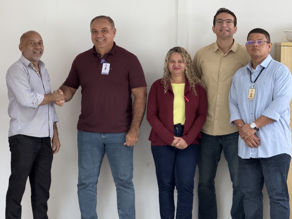 Grupo de cinco pessoas posando para foto em ambiente interno, alinhadas lado a lado. À esquerda, dois homens apertam as mãos e sorriem. No centro, uma mulher com cabelos cacheados e roupa em tons de vermelho e amarelo. À direita, dois homens em pé, um deles usando crachá. Todos estão voltados para a câmera, em clima formal e amistoso.